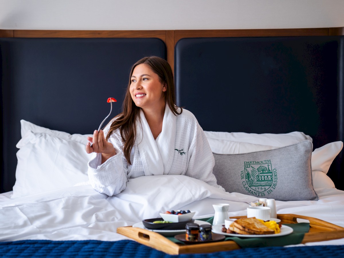 A woman in a robe enjoys breakfast in bed with a tray of food, smiling and holding a fork with a strawberry on it.
