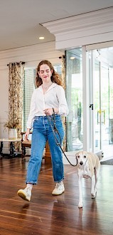 A person walks a dog through a bright, modern lobby with wooden floors and white walls, towards a glass entrance.