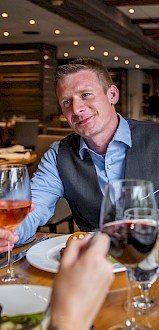 A man in a blue shirt and vest enjoys a meal at a restaurant, holding a glass of wine, with another person.