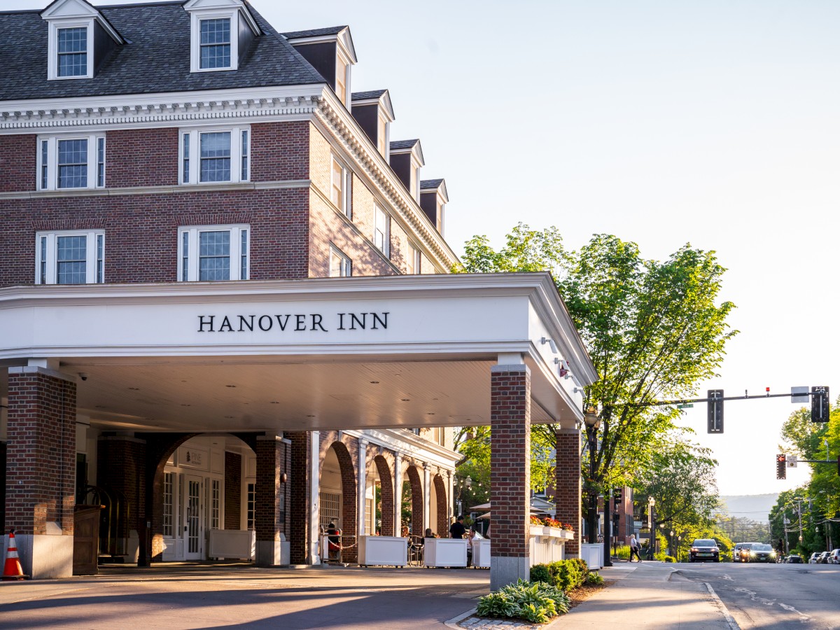 Street view of Hanover Inn with a large canopy at the entrance, brick facade, and greenery around. Traffic lights are visible further down the road.