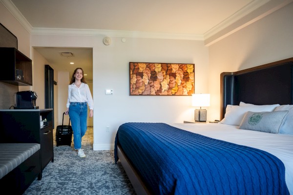 A person entering a modern hotel room with a large bed, artwork, and a nightstand with a lamp.