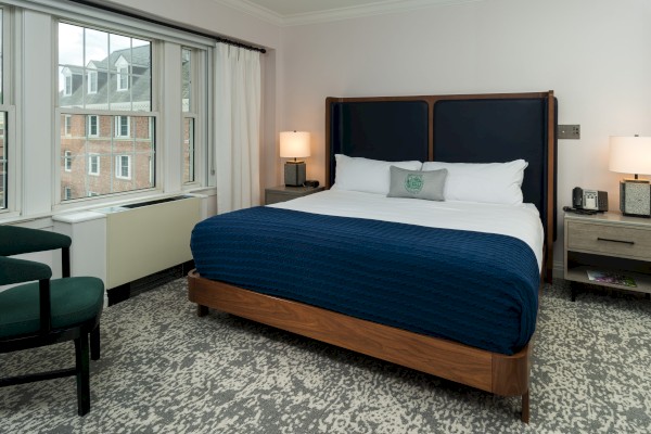 A modern hotel room with a king-size bed, blue blanket, nightstands, lamps, a large window with a view, and chairs in the corner, ending the sentence.