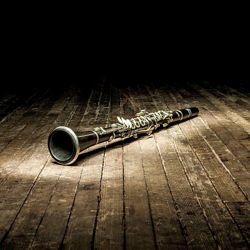 A clarinet lies on a wooden floor, spotlighted in a dimly lit setting, highlighting its polished surface and intricate details.