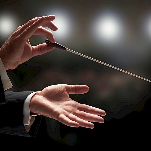 A conductor's hands holding a baton, captured mid-performance with blurred lights in the background, suggesting a musical setting.