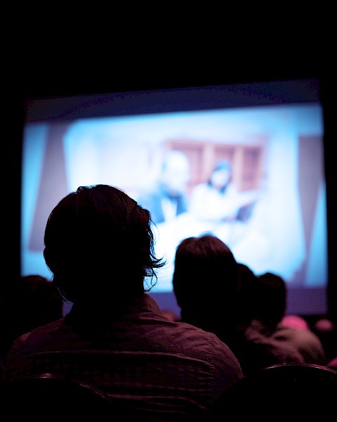 People sitting in a dark room watching a large screen displaying a blurred image, possibly a movie or presentation, in a cinema or theater.