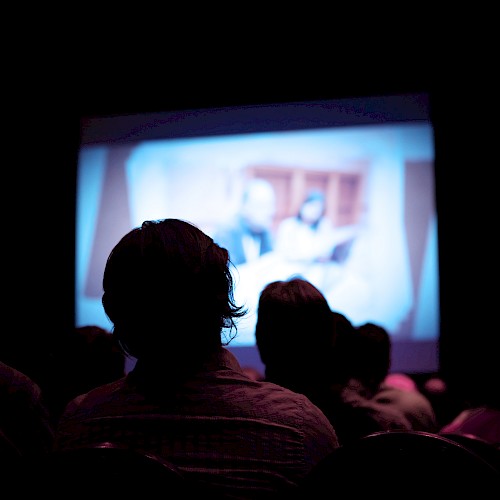 People sitting in a dark room watching a large screen displaying a blurred image, possibly a movie or presentation, in a cinema or theater.