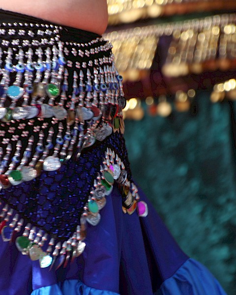 The image shows a vibrant, beaded belly dance hip scarf with colorful sequins and fringe details on a richly colored skirt.