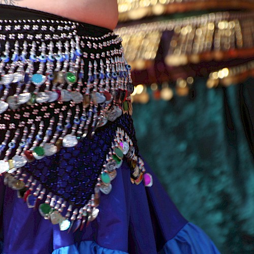 The image shows a vibrant, beaded belly dance hip scarf with colorful sequins and fringe details on a richly colored skirt.