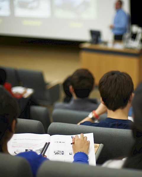 Students are sitting in a lecture hall taking notes, with a lecturer presenting at the front of the room.