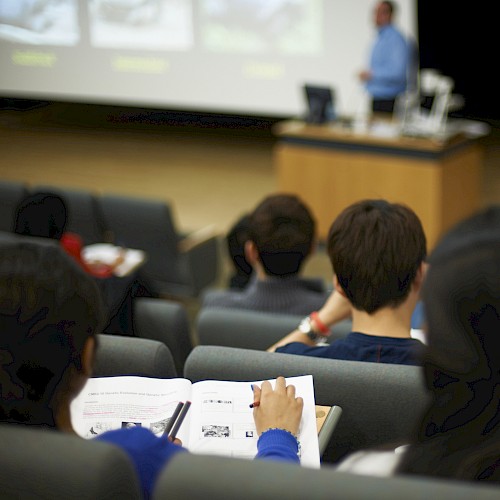 Students are sitting in a lecture hall taking notes, with a lecturer presenting at the front of the room.