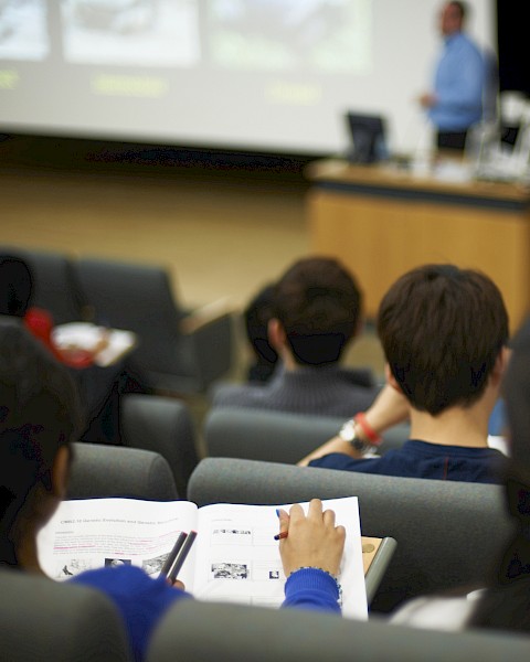 The image shows students sitting in a lecture hall, taking notes while a lecturer presents at the front.