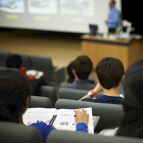 The image shows students sitting in a lecture hall, taking notes while a lecturer presents at the front.