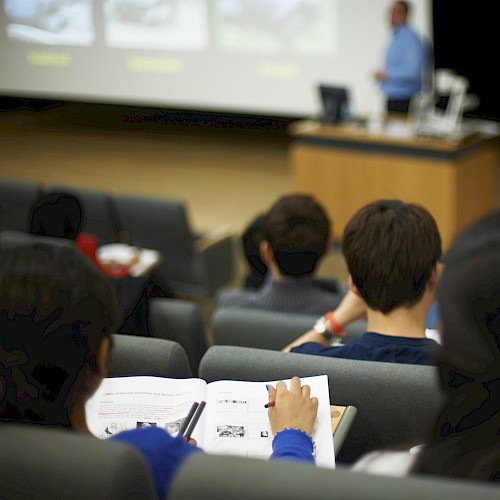 Students are sitting in a lecture hall, attentively watching a presentation on a screen, with notes and materials in hand.