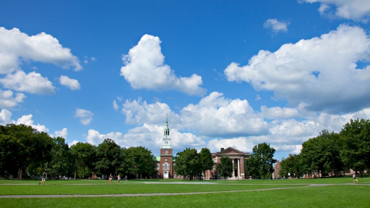 A grassy area with scattered pedestrians, surrounded by trees and featuring a tall clock tower and classical building under a blue sky.