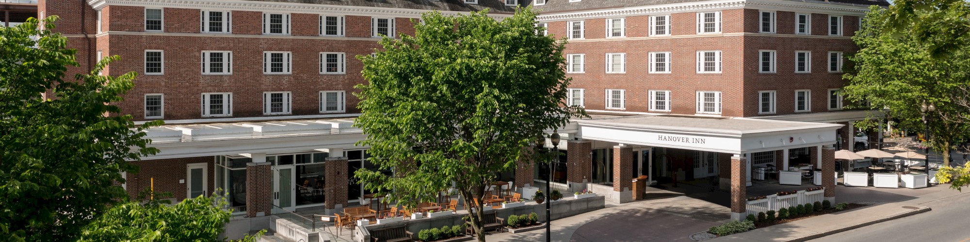 A large red-brick building with multiple windows, surrounded by trees and a road in front, with clear blue skies above.