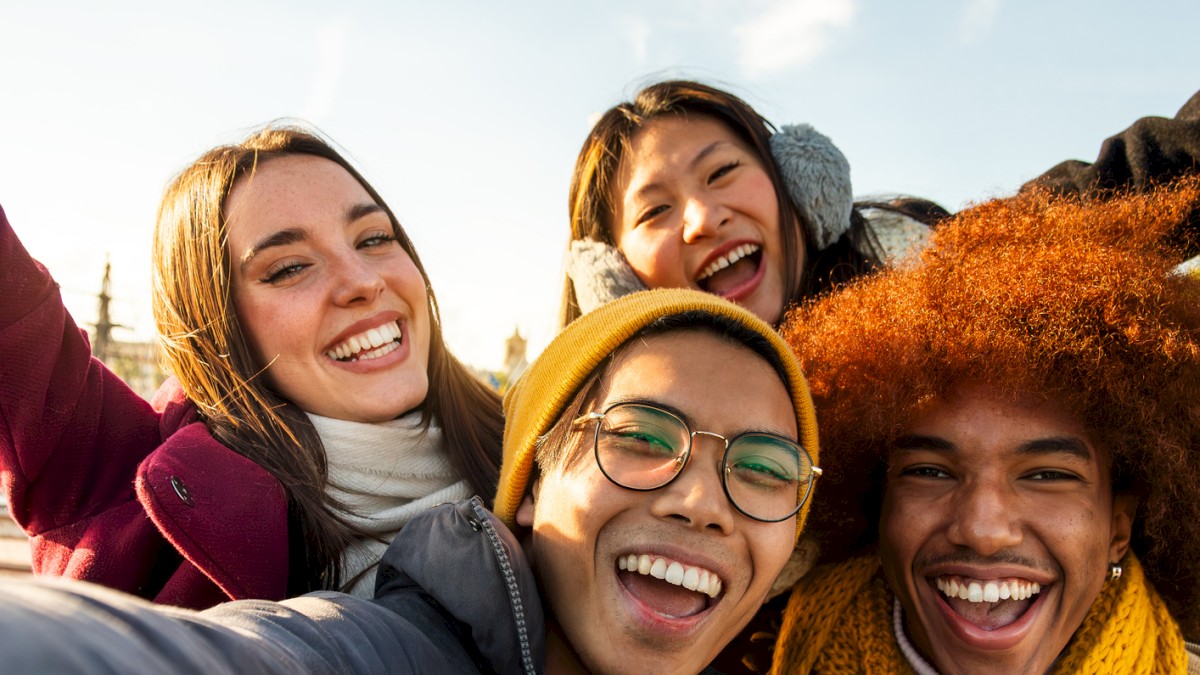 Four people are taking a cheerful group selfie outdoors, with a bright sky and a bird flying in the background.