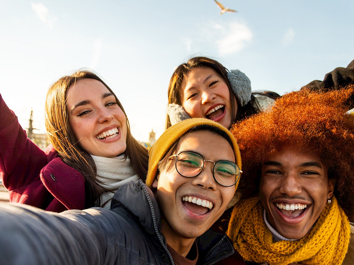 Four people are taking a cheerful group selfie outdoors, with a bright sky and a bird flying in the background.