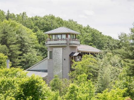 A multi-level building with a tower is surrounded by lush green trees and foliage under a partly cloudy sky.