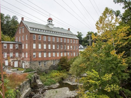 A large brick building with many windows is beside a small river and surrounded by trees. Power lines are visible in the sky.