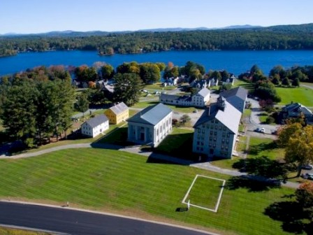 An aerial view of a campus near a lake, featuring several large buildings, expansive green lawns, and surrounding trees and hills.