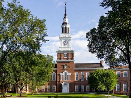 A historic brick building with a tall central clock tower, surrounded by trees and greenery under a blue sky.