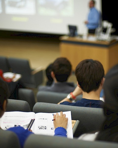 Students in a lecture hall take notes while a presenter speaks at a podium, with a projected presentation screen in the background.