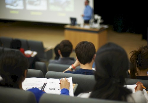 Students in a lecture hall take notes while a presenter speaks at a podium, with a projected presentation screen in the background.