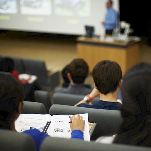 Students in a lecture hall take notes while a presenter speaks at a podium, with a projected presentation screen in the background.