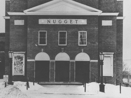 A black and white photo of the Nugget building, featuring three entrance arches, a sign, and snow-covered ground in front.