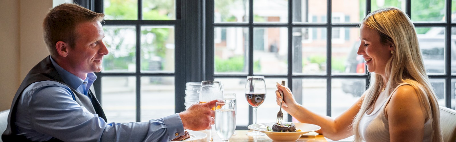 A man and woman are dining at a table in a restaurant, raising their wine glasses for a toast, while smiling at each other.
