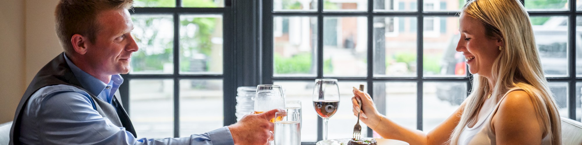 A man and woman are dining at a table in a restaurant, raising their wine glasses for a toast, while smiling at each other.