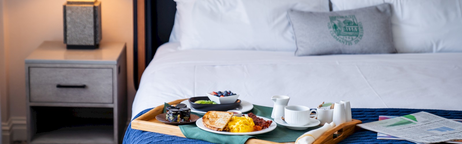 A neatly made bed with a breakfast tray, including food, tea, and papers, sits under a lamp on a bedside table.