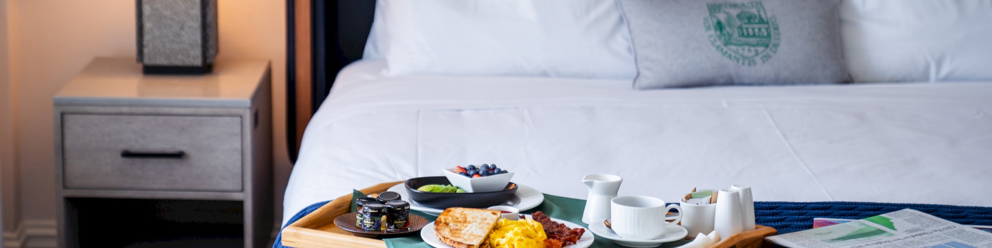 A neatly made bed with a breakfast tray, including food, tea, and papers, sits under a lamp on a bedside table.