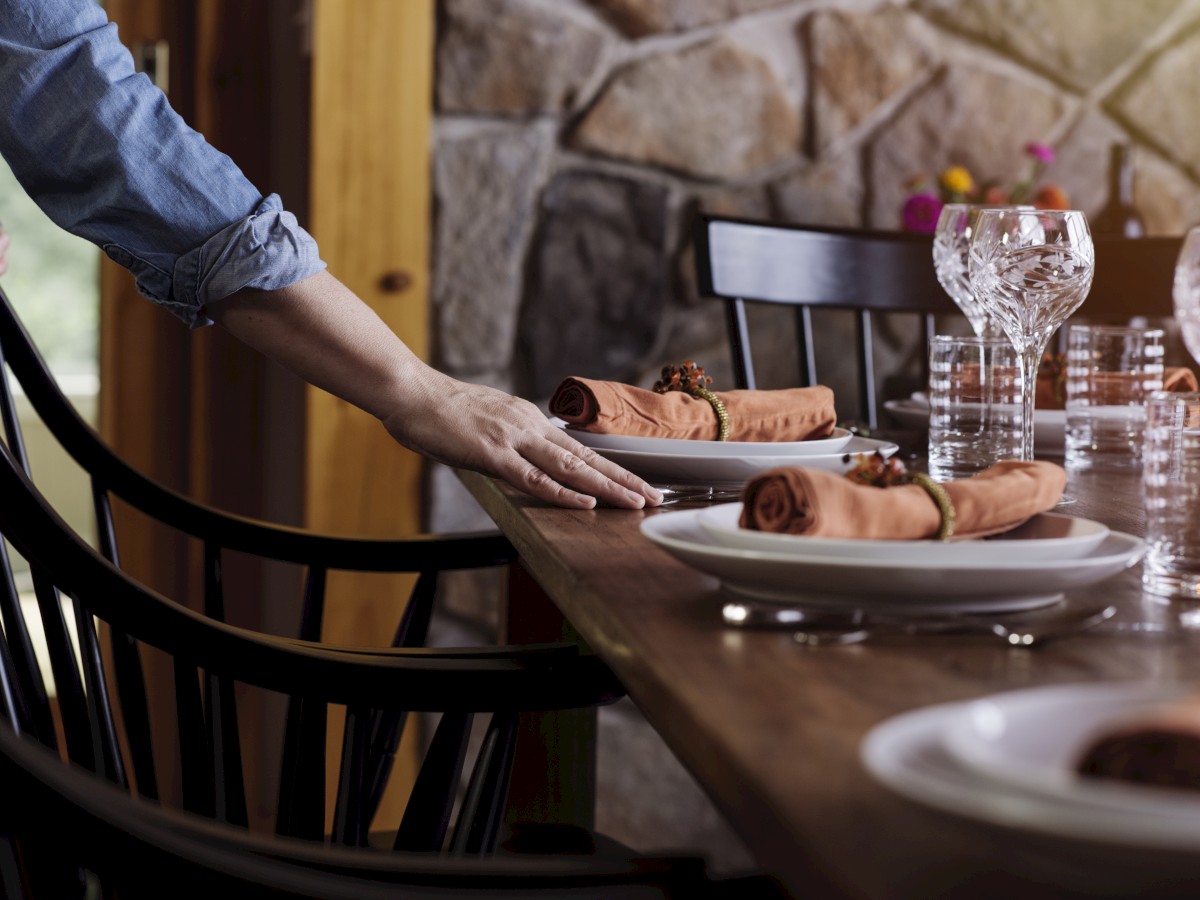 A person is setting a dining table with plates, folded napkins, and glasses, in a room with stone walls and wooden chairs.