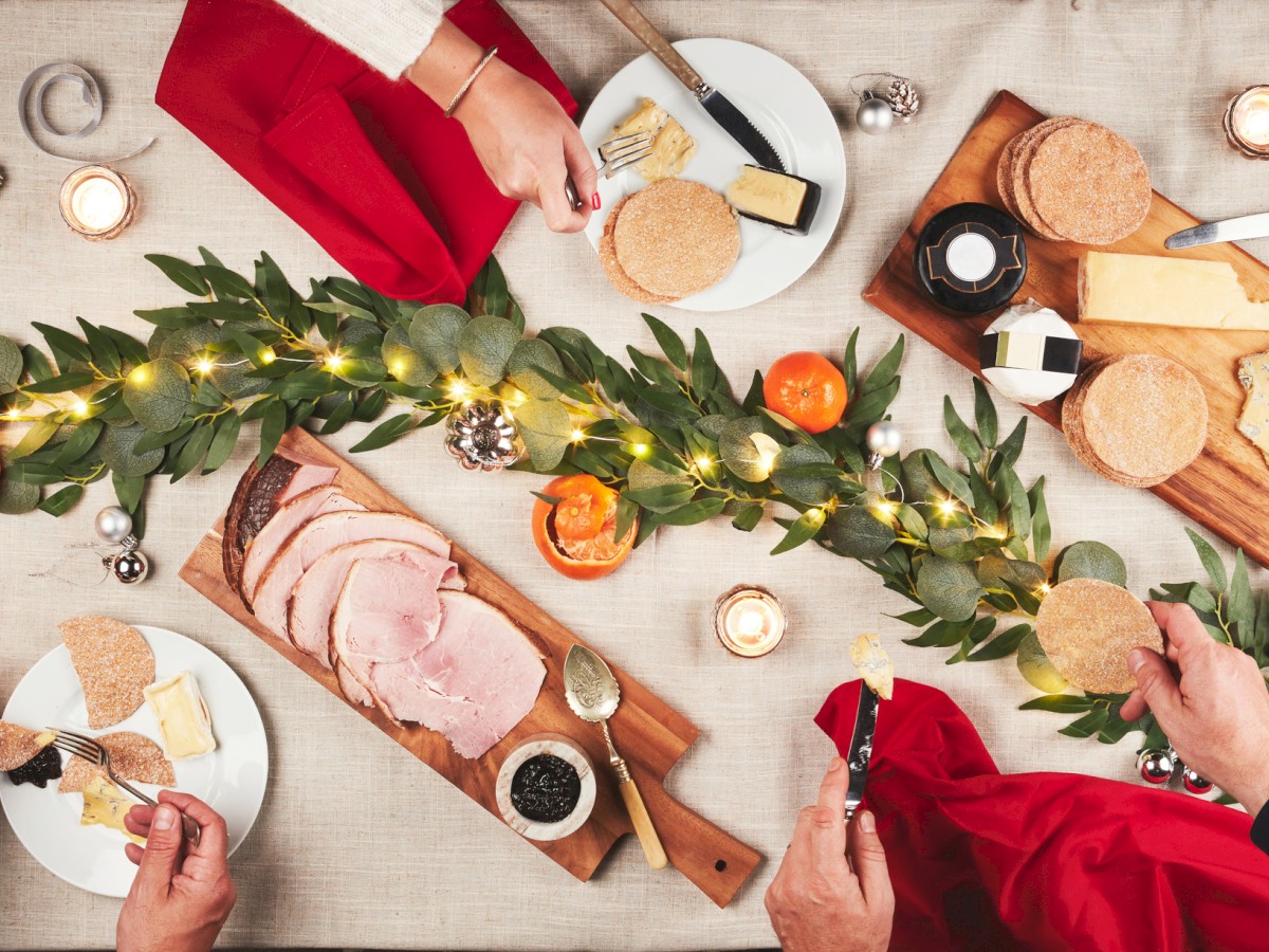 A festive table setting with ham, cheese, crackers, and fruits, adorned with greenery and lights. Hands are seen serving food around it.