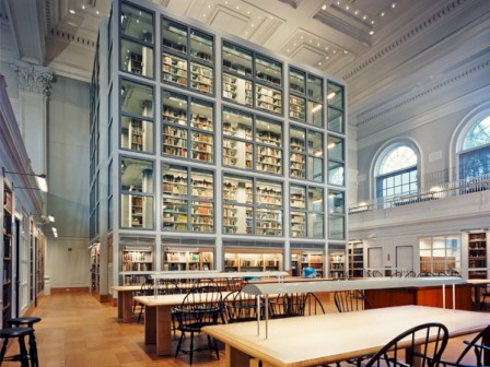 The image shows an elegant library with high ceilings, featuring a large glass-enclosed wall of books and study tables with chairs below.