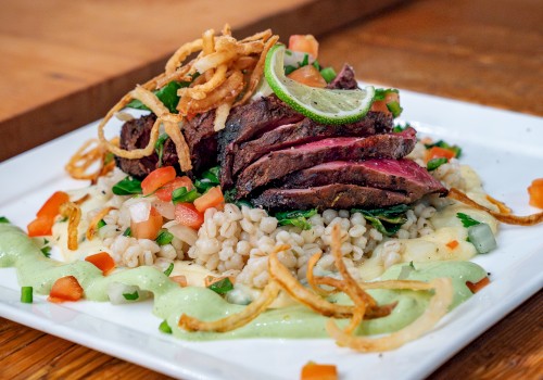 The image shows a gourmet dish with sliced steak on a bed of grains, garnished with fried onions, greens, and a creamy sauce on a square plate.