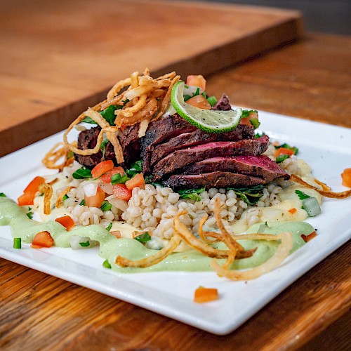 The image shows a gourmet dish with sliced steak on a bed of grains, garnished with fried onions, greens, and a creamy sauce on a square plate.