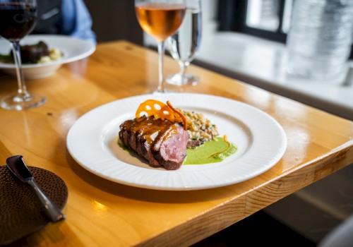 A plate with sliced steak, sauce, and garnish, sitting on a wooden table with wine glasses in the background.