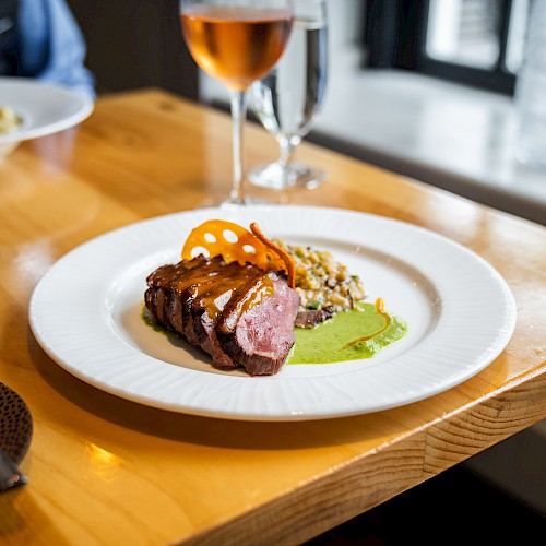 A plate with sliced steak, sauce, and garnish, sitting on a wooden table with wine glasses in the background.