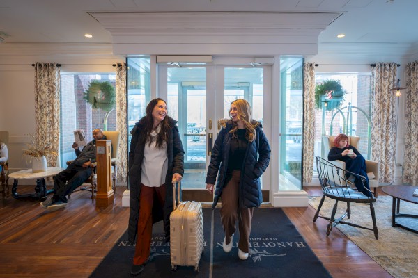 Two women with suitcases stand at a hotel lobby entrance, smiling and chatting, while guests relax on chairs around and through glass doors.