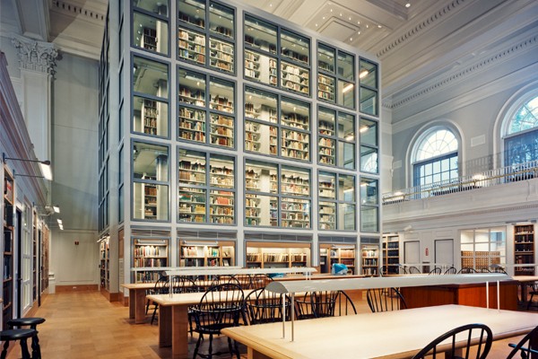 The image shows a library with tall, glass-enclosed bookshelves, wooden tables, and chairs in a spacious, well-lit room with high ceilings.