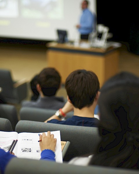 Students in a lecture hall, sitting on chairs, viewing a presentation on a screen while taking notes during a class.