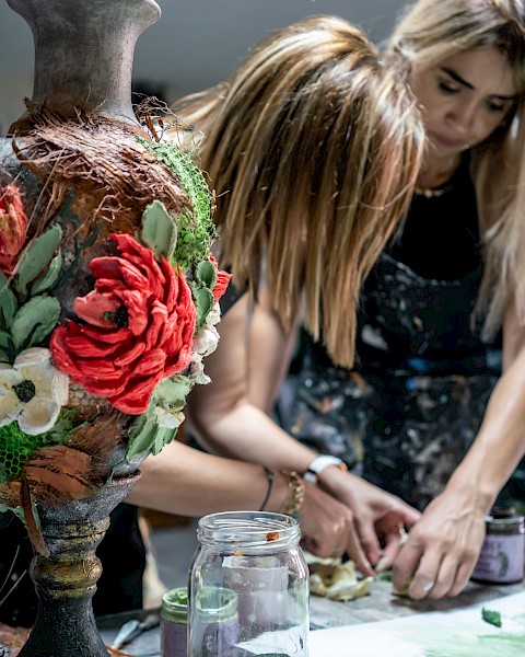 Three people crafting at a table with a decorative vase in the foreground, featuring large, colorful flowers.