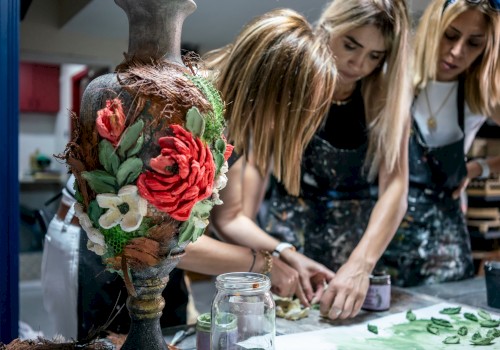 Three people crafting at a table with a decorative vase in the foreground, featuring large, colorful flowers.