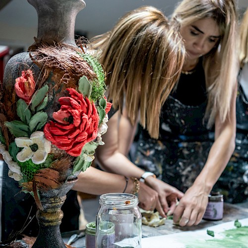 Three people crafting at a table with a decorative vase in the foreground, featuring large, colorful flowers.