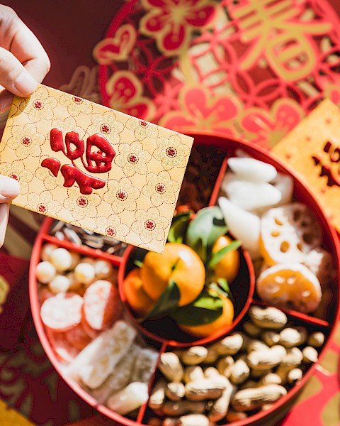 A person holds a red envelope above a tray of Lunar New Year treats, including nuts, candied fruits, and tangerines, with red decorations.