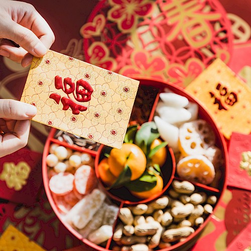 A person holds a red envelope above a tray of Lunar New Year treats, including nuts, candied fruits, and tangerines, with red decorations.