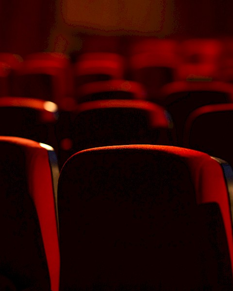 The image shows a dimly lit theater or cinema with rows of empty red seats, creating an intimate and quiet atmosphere.