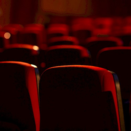 The image shows a dimly lit theater or cinema with rows of empty red seats, creating an intimate and quiet atmosphere.